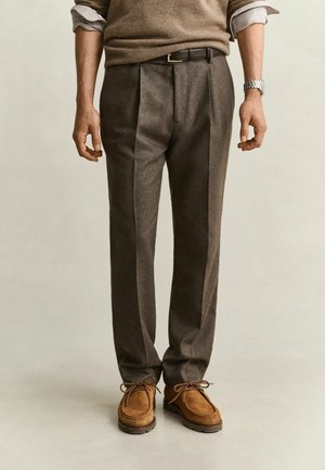 Man draagt bruine geplooide broek, lichtbruine suède veterschoenen, een bruine trui over een opgerolde witte overhemd en een zilveren polshorloge.