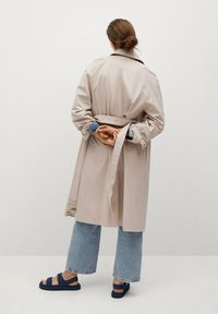 Beige trenchcoat met ceintuur in de taille, lichtgewicht stof, brede mouwen, gecombineerd met blauwe flared jeans en donkere sandalen. Minimalistisch ontwerp.