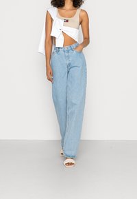 Crop top beige con logo, felpa bianca oversize drappeggiata e jeans a gamba larga di un blu chiaro. Sandali bianchi semplici con cinturini completano l'outfit.