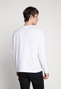 Tricou alb cu mânecă lungă, realizat din bumbac. Are gât rotund și tiv drept, prezentat din spate, fără modele sau grafică vizibile.