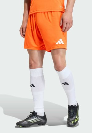 Mann trägt leuchtend orangefarbene Sportsshorts, weiße kniehohe Socken mit schwarzem Logo und schwarze Fußballschuhe mit neongrünen Akzenten.