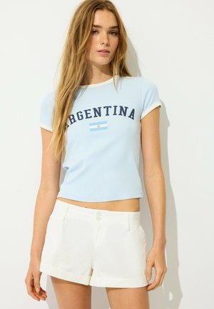 Młoda kobieta z długimi włosami ubrana w jasnoniebieski krótki top z "Argentina" i flagą, w połączeniu z białymi, wysokimi szortami.