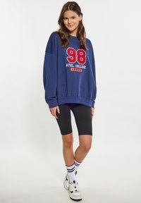 Felpa navy con grafica rossa e bianca "98 ATHL. COLLEGE" sul davanti, abbinata a pantaloni da ciclismo neri e sneakers bianche con accenti neri.