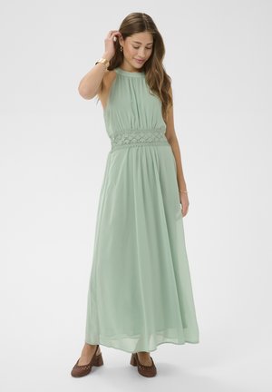 Žena v bezrukávových svetlozelených maxi šatách s kvetinovým pásom v páse, v hnedej obuvi na podpätku a so zlatými šperkami, pózujúca s jednou rukou dotýkajúcou sa vlasov.