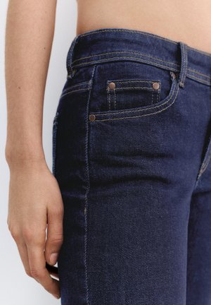 Henkilön vasen puoli, jossa päällä tummansiniset farkut, joista näkyy etutasku ja kolikkotasku, käsi rentona lantion vieressä, paljas keskivartalo.