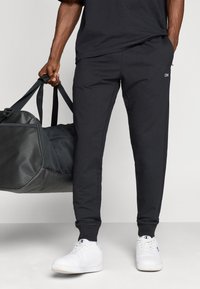 Czarne spodnie dresowe z elastycznymi mankietami, wykonane z miękkiego materiału. Udekorowane małym logo na udzie. W zestawie z czarną torbą na siłownię i białymi sneakersami.