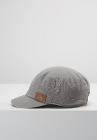 Gorra de tela gris con visera plana, material texturizado y una etiqueta de cuero marrón con un logotipo; incluye dos detalles de botones en el lado.