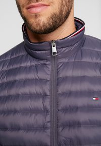 Giacca imbottita da uomo in tessuto viola scuro con cuciture orizzontali, colletto alto e zip argentata. Presenta un piccolo logo in rosso, bianco e blu.