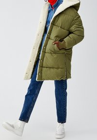 Abrigo acolchado verde con forro de piel sintética blanca, capucha y bolsillos con cremallera, superpuesto a una chaqueta de mezclilla y pantalones vaqueros azules. Zapatillas blancas.