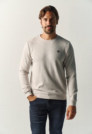 Hombre con barba y cabello corto que lleva una sudadera beige clara y vaqueros oscuros, de pie con una mano en el bolsillo sobre un fondo liso.