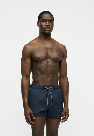 Veltrent mørkhudet mann med kort hår står vendt mot oss, iført marineblå svømmeshorts med grå snor på en ensfarget lys bakgrunn.