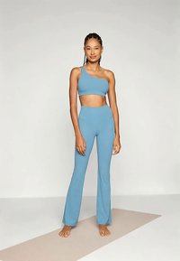 Mlada ženska stoji bosa na bež jogijski blazini, oblečena v svetlo modro enostransko kratko majico in ujemajoče se široke hlače, ob ozadju brez vzorca.