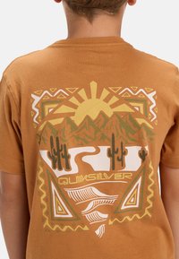 Hnedé bavlnené tričko s farebnou potlačou hôr, kaktusov, slnka a geometrických vzorov, so zvýrazneným textom "Quiksilver" na spodnej časti.