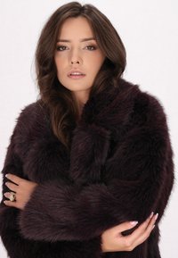 Plysset bordeaux faux fur frakke med høj krave, brede ærmer og en blød tekstur. Modellen har hænderne krydset, iført en ring på den ene hånd.