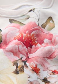 Povečan prikaz tkanine z detajlnim rožnatim in belim cvetličnim potiskom, ki prikazuje velike cvetne lističe in zelene liste na svetli podlagi.