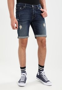 Modèle masculin portant un short en denim déchiré et des baskets montantes navy avec des lacets blancs, sur fond blanc uni.