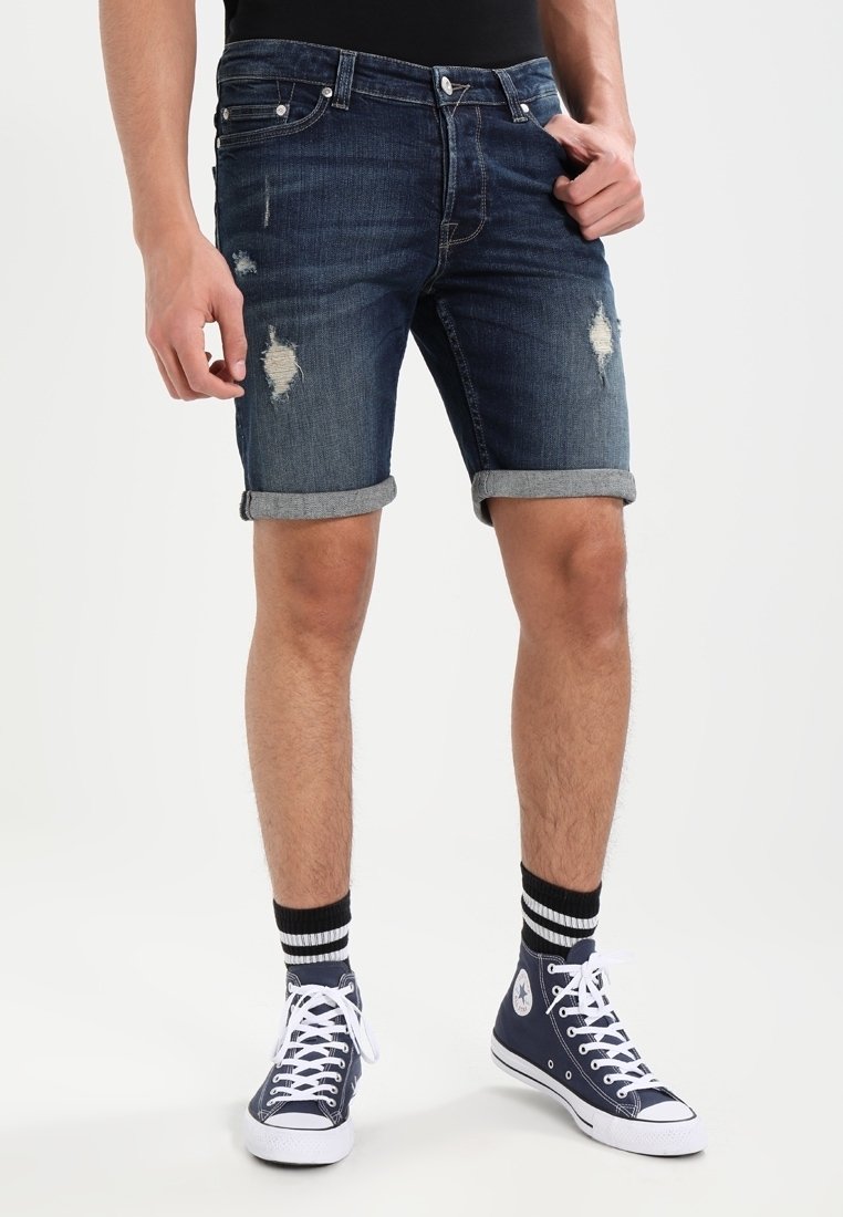 Modèle masculin portant un short en denim déchiré et des baskets montantes navy avec des lacets blancs, sur fond blanc uni.