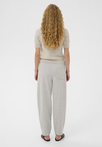 Femeie cu păr lung și creț, îmbrăcată într-un pulover bej cu mânecă scurtă, pantaloni largi gri deschis cu buzunar pe spate și pantofi negri slip-on.