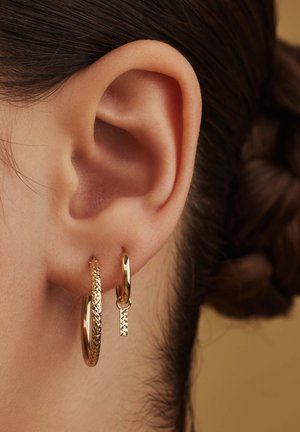 Gold hoop earrings, one textured and one smooth, hanging from an ear. The textured earring has a thin geometric charm attached.