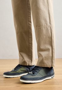 Zapatillas de textil navy con acentos amarillos, que presentan una parte superior de malla, suelas de goma blancas y cordones negros, combinadas con pantalones beige claro.