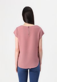 Krátký rukáv, růžový top z lehkého materiálu s jemnou texturou. Obsahuje zip na zádech a zakulacený lem. V kombinaci s modrými džíny.