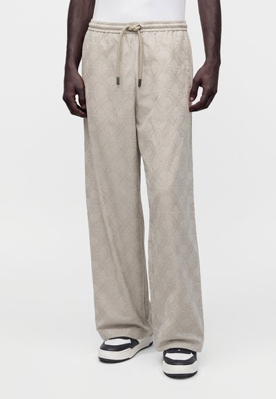Pantalon beige à jambes larges avec un motif géométrique subtil, taille avec cordon, porté avec des chaussures slip-on noires et blanches sur une personne à la peau foncée.