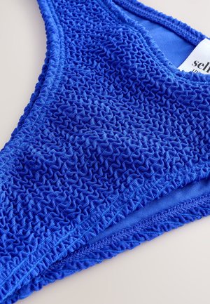 Kék bikini alsó texturált, fodros anyaggal. Simított szélekkel és varrás nélküli dizájnnal rendelkezik, kiemelve az élénk, egyszínű színt.