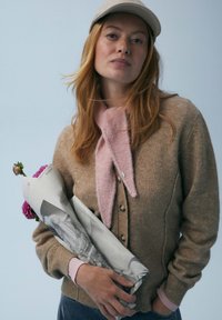 Cardigan in lana beige con bottoni di legno, indossato sopra una camicia a maniche lunghe rosa. Una sciarpa rosa chiaro avvolge il collo, sorreggendo un giornale arrotolato e dei fiori.