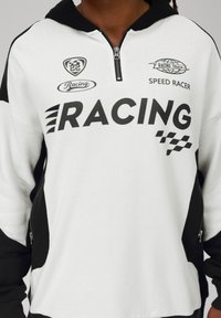 Balts un melns kapucētais džemperis ar rāvējslēdzēju apkaklē, ar vārdiem "RACING" treknrakstā melnā krāsā un dažādiem logotipiem drukātiem uz priekšpuses.