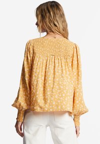 Blouse jaune avec un motif floral blanc, corsage froncé et manches bouffantes longues. Le tissu semble léger avec une texture douce.