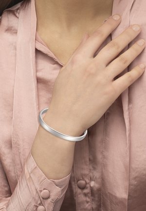 Sølvfarget metallisk armring på en hånd, med en jevn, rund design. Hånden hviler mot en lys rosa silke bluse.