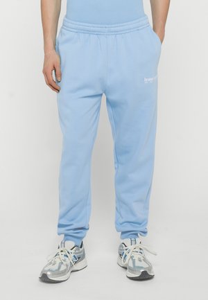 Persona que lleva puestos unos pantalones deportivos azul claro con un pequeño logo blanco en el muslo izquierdo y zapatillas deportivas blancas, azules y grises, de pie con las manos en los bolsillos.