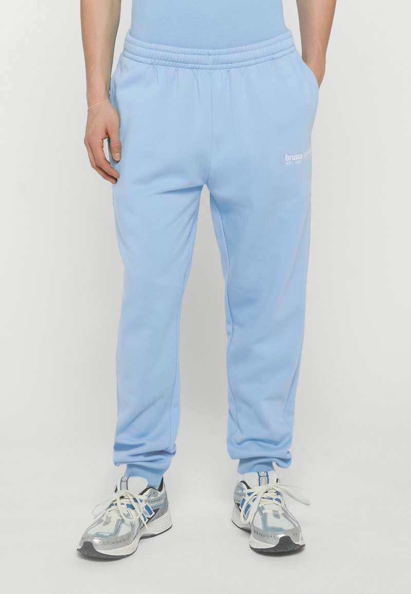Personne portant un pantalon de survêtement bleu clair avec un petit logo blanc sur la cuisse gauche et des chaussures de sport blanches, bleues et grises, debout les mains dans les poches.