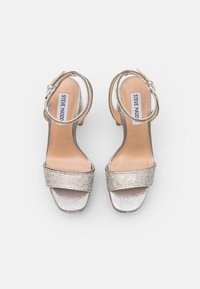 Sandalen mit hohen Absätzen aus silbernem Stoff, ausgestattet mit einem breitbandigen Vorderteil, verziert mit Strasssteinen, und einem verstellbaren Knöchelriemen.