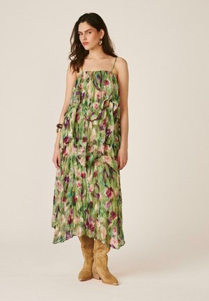 Vrouw draagt een groene, bloemenprint mouwloze midijurk met ruches en beige suède cowboylaarzen, staand tegen een effen achtergrond.