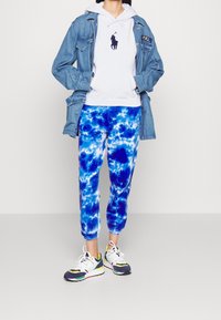 Jeanjacka över en vit hoodie med blå batikmönstrade joggers och flerfärgade skor. Viktiga detaljer inkluderar fickor och ribbad mudd.