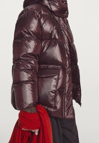 Donkerrode donsjas met capuchon, glanzende textuur en zichtbare stiksels. Voorzien van grote zakken en een aansluitend silhouet. Houdt een rode sjaal vast.