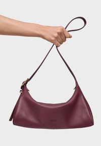 Mano sosteniendo un bolso de hombro de cuero granate con una sola correa y herrajes de tono dorado, con el nombre de la marca TOUS grabado en relieve.