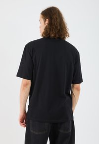 Black cotton t-shirt with short sleeves, crew neck, and a relaxed fit, viewed from the back. Notable for its simple design and minimal detailing.