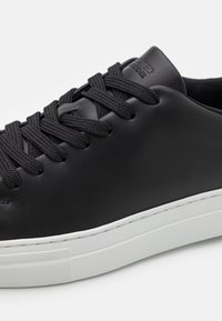 Mustat nahkasneakerit, joissa on teksturoidun yläosan, pyöreä kärki ja tasainen valkoinen kumipohja. Varustettu mustilla kudotuilla nauhoilla ja hienovaraisella brändin painatuksella.