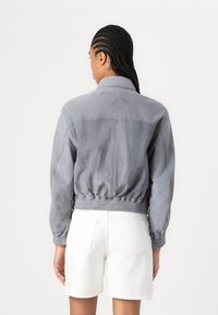 Giacca bomber in suede grigio con design corto, vita elastica e maniche lunghe. Visibili pantaloni shorts in denim bianchi, sfondo semplice.