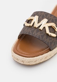 Sandale marron en cuir à enfiler avec semelle en jute tressée, dotée d’une bride foncée à motifs et d’un détail décoratif doré avec le logo « MK ».