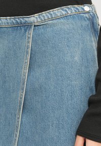 Jupe en denim bleu clair avec couture verticale et bouton argenté à la taille, portée sur un haut noir côtelé à manches longues.