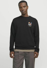 Sudadera negra de cuello redondo, con un parche gráfico colorido en el pecho. Tejido suave y texturizado; corte relajado. Combinada con pantalones en tonos neutros.