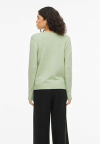 Femme aux cheveux bouclés vue de dos, portant un pull vert clair et un pantalon noir à jambes larges, sur un fond blanc.