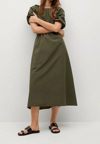 Olivengrünes Baumwollkleid mit Puffärmeln und elastischem Bund, das eine lange A-Linie-Silhouette hat, kombiniert mit schwarzen Rutsch-Sandalen.