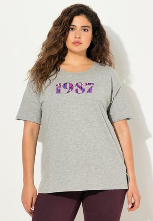 T-shirt gris à manches courtes en tissu doux, arborant l'année "1987" en impression florale violette sur le devant.
