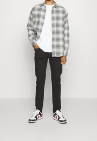 Man som bär en grå ruta skjorta med knappar över en vit t-shirt, svarta byxor och vita sneakers med röda och svarta detaljer, står med en hand i fickan.