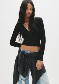 Schwarzes Langarmshirt mit geripptem Muster und V-Ausschnitt, kombiniert mit hellblauen, distressed Jeans und einem gebundenen schwarzen Hemd um die Taille.