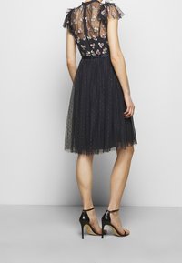 Robe en tulle noir avec broderies florales sur un haut transparent, manches volantes et corsage ajusté. Associée à des sandales à talons hauts noires.